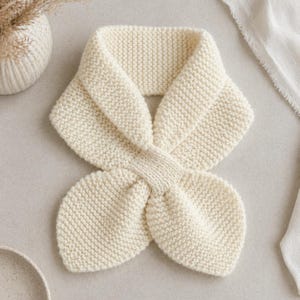 Könnte beinhalten: Cremefarbener Strickschal mit einem einzigartigen, geschlungenen Design. Der Schal ist aus weichem, strukturiertem Garn gefertigt und hat eine unverwechselbare Form, perfekt, um einen Hauch von Wärme und Stil zu verleihen. Ein gemütliches Accessoire.