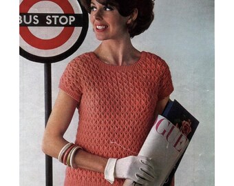 May include: A woman in a coral-coloured, short-sleeved knitted top with a diamond pattern. She holds a magazine with "Vogue" visible. A bus stop sign is in the background.