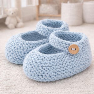Könnte beinhalten: Ein Paar hellblaue gestrickte Babyschuhe. Die Schuhe haben einen Mary-Jane-Stil mit Riemen und einem Holzknopfverschluss. Die Schuhe sind aus weichem, strukturiertem Garn gefertigt und für Babys geeignet.