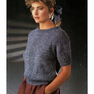 May include: A blue knitted short-sleeved jumper with a textured pattern. The jumper has a round neckline and a slightly fitted shape. The model is wearing a red patterned skirt and large earrings.