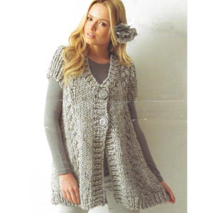 Könnte beinhalten: Hellgraue, kurzärmelige Strickjacke mit V-Ausschnitt und zwei großen, dekorativen Knöpfen. Die Strickjacke hat ein strukturiertes Strickmuster und eine leicht ausgestellte Form. Das Kleidungsstück ist etwa 76 cm lang.