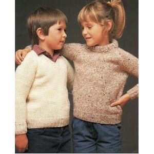 May include: Two children wearing knitted sweaters. One child is wearing a cream-colored V-neck sweater over a plaid shirt. The other child is wearing a speckled brown sweater with a high neckline. Both are wearing blue jeans.