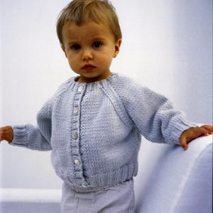 May include: A light blue knitted cardigan with button closures. The baby sweater has long sleeves and a ribbed hem and cuffs. The child is wearing light gray pants. The background is a plain white.