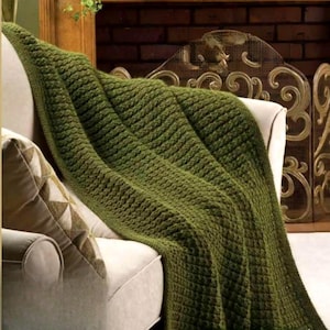 May include: An olive green knitted throw blanket draped over a cream armchair. The blanket features a textured, woven pattern. A decorative pillow with a geometric design rests on the chair. The background includes a fireplace and decorative screen.