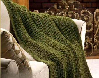 May include: An olive green knitted throw blanket draped over a cream armchair. The blanket features a textured, woven pattern. A decorative pillow with a geometric design rests on the chair. The background includes a fireplace and decorative screen.