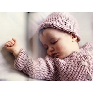May include: A sleeping baby wearing a knitted pink hat and cardigan. The cardigan has three white buttons. The baby's arm is extended, and the baby is lying on a white surface.