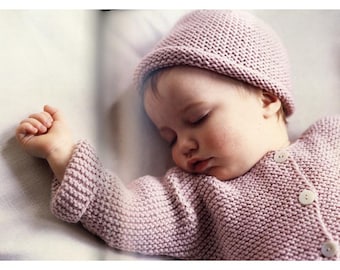 May include: A sleeping baby wearing a knitted pink hat and sweater. The sweater has three white buttons. The baby's arm is extended, and the baby is lying on a white surface.