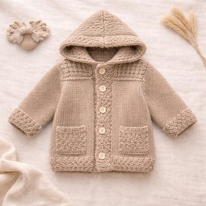 May include: A tan knitted baby jacket with a hood, button closure, and two front pockets. The jacket has a textured knit pattern on the cuffs, pockets, and hood. A matching headband and dried wheat stalks are also visible.