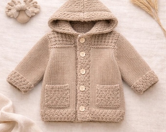 May include: A tan knitted baby jacket with a hood, button closure, and two front pockets. The jacket has a textured knit pattern on the cuffs, pockets, and hood. A matching headband and dried wheat stalks are also visible.
