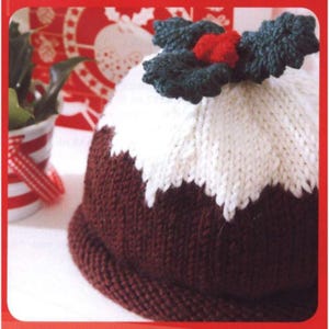 Könnte beinhalten: Gestrickte Weihnachtspudding-Mütze mit dunkelbraunem Boden, weißer Glasur und grünen Stechpalmenblättern mit roten Beeren. Ein festliches Accessoire für die Weihnachtszeit.
