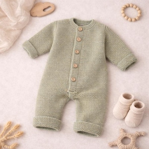 Peut inclure: Un barboteuse de bébé tricotée vert clair avec des manches longues et des boutons en bois. La barboteuse a des poignets repliés sur les manches et les jambes. Une paire de chaussettes de bébé crème et des jouets en bois sont également dans l'image.