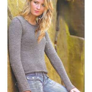 May include: A long-sleeved, gray knit sweater with a crew neck. The sweater is paired with blue denim jeans. The model is leaning against a textured, yellow-toned wall. The sweater has ribbed cuffs and hem.