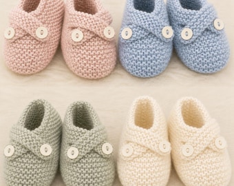 May include: A collection of six knitted baby booties in pastel colors: pink, blue, green, and cream. Each pair features a cross-over design secured with a white button. The booties are arranged on a soft, light-colored surface.