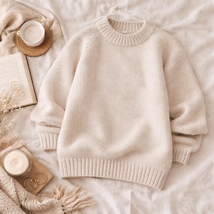Könnte beinhalten: Cremefarbener Strickpullover mit geripptem Rundhalsausschnitt und Bündchen. Der Pullover wird auf einem Bett mit einem Buch, einer Tasse Kaffee, einer Kerze und goldenen Creolen präsentiert.
