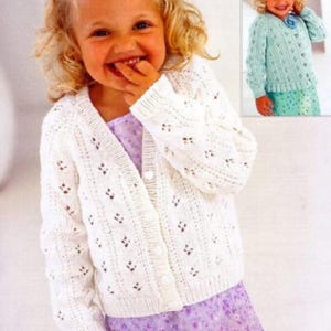 Op de afbeelding: Een witte cardigan met open voorkant en een delicaat open breipatroon. De trui heeft lange mouwen en wordt met knopen gesloten. Het ontwerp bevat kleine, ruitvormige uitsparingen. Het wordt gedragen over een paarse jurk.