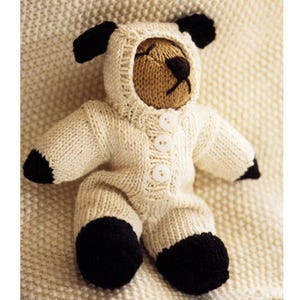 May include: A knitted teddy bear wearing a white hooded jumpsuit with black detailing on the ears, paws, and feet. The jumpsuit features three white buttons. The bear's face is tan with a black nose and embroidered features.