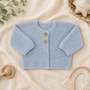 May include: A light blue long-sleeve knit cardigan featuring a round neckline and two small, round, light brown wooden buttons centred on the front placket. The garment is constructed with a consistent garter stitch texture throughout.