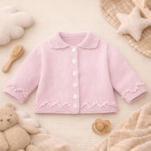 May include: A pale pink knitted baby cardigan with a rounded collar and button fastening. The long sleeves and hem have a decorative pattern. Baby accessories, including a teddy bear and dummy, surround the cardigan.