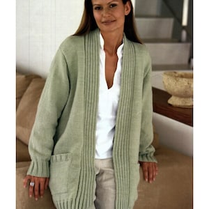 May include: A light green open-front cardigan with a ribbed design on the front and pockets. The cardigan is worn over a white blouse and beige pants. The sleeves have ribbed cuffs. The cardigan is made of a soft, knitted material.