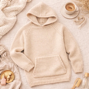 May include: A cream-coloured knitted hoodie with a kangaroo pocket. The hoodie has a hood and long sleeves. The image also includes a cup of coffee, a small dish with beauty products, and a wooden hairbrush.