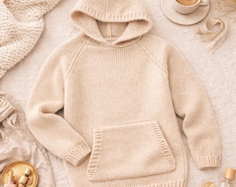 May include: A cream-colored knitted hoodie with a kangaroo pocket. The hoodie has a hood and long sleeves. The image also includes a cup of coffee, a small dish with beauty products, and a wooden hairbrush.
