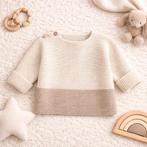 Può includere: Maglione per neonati color crema e beige con scollo tondo e maniche lunghe. Il maglione ha bottoni in legno sulla spalla e polsini a coste. Un giocattolo di legno, un cuscino a forma di stella e un orsacchiotto sono anche nell'immagine.