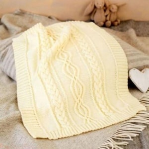 May include: A cream-colored knitted blanket with a cable knit design. The blanket has a textured appearance with a ribbed border. The blanket is folded over a pillow and blanket. A wooden heart and stuffed animals are in the background.