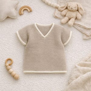 May include: A beige knitted baby shirt with a V-neck and short sleeves, trimmed with white. The shirt is surrounded by a plush toy bunny, a wooden rainbow, and other baby items. The shirt is approximately 10 inches long.