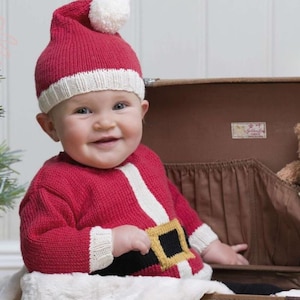 May include: A baby in a red knitted Santa Claus outfit, featuring a hat with a white bobble and a sweater with a white belt and gold buckle. The baby smiles, seated in a brown suitcase.