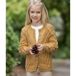 Può includere: Un cardigan giallo senape a maglia a trecce con bottoni in legno. Il cardigan è indossato sopra una camicia bianca. La persona tiene una pigna. Lo sfondo è verde sfocato.