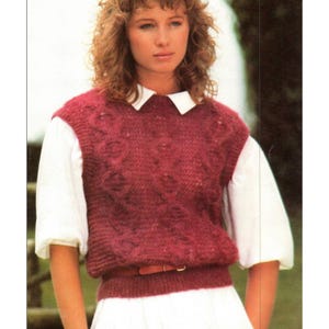 May include: A burgundy knitted vest with a textured pattern is worn over a white collared shirt. A brown belt cinches the waist. The model is wearing a white skirt. The image is a vintage fashion photograph.