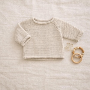 May include: A cream-coloured knitted baby sweater with long sleeves and ribbed cuffs and hem. A wooden teething ring and a small sprig of dried flowers are next to the sweater. The sweater is made of a soft, textured yarn.