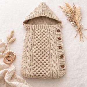 以下が含まれることがあります： ベージュ色のニットベビー寝袋。フードと木製のボタンが付いています。寝袋は、側面にケーブルニットのデザイン、中央にダイヤモンドパターンが特徴です。木製の歯固めとドライフラワーも画像にあります。