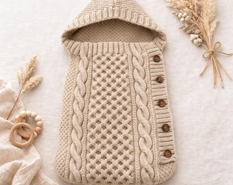 May include: A beige knitted baby sleeping bag with a hood and wooden buttons. The sleeping bag features a cable knit design on the sides and a diamond pattern in the center. A wooden teething ring and dried flowers are also in the image.