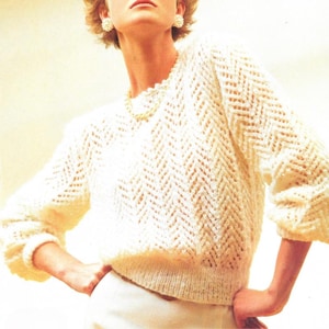 Könnte beinhalten: Cremefarbener Pullover mit offenem Gewebe und Zickzackmuster. Der Pullover hat einen Rundhalsausschnitt und Dreiviertelärmel. Der Saum und die Manschetten sind gerippt. Das Model trägt eine Perlenkette und Ohrringe.