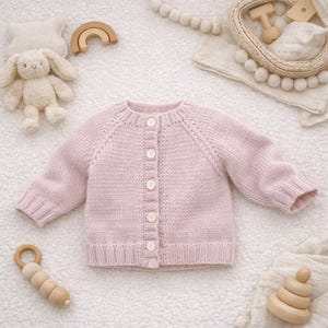 Può includere: Un cardigan rosa chiaro lavorato a maglia per neonati, con maniche lunghe e chiusura con bottoni. Il maglione è realizzato in filato morbido e ha orlo e polsini a coste. Lo sfondo include un coniglietto di peluche, giocattoli di legno e una ghirlanda decorativa.