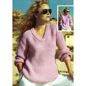 Può includere: Un maglione rosa chiaro con scollo a V e dettagli a coste. Il maglione ha maniche lunghe con polsini arricciati. L'immagine include anche una piccola immagine dello stesso maglione. La modella indossa occhiali da sole e pantaloni bianchi.