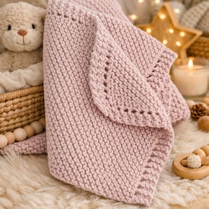 Puede incluir: Una suave manta de bebé de punto rosa con un borde decorativo. La manta está doblada, mostrando la textura del tejido. Un osito de peluche y juguetes de madera están en el fondo, sugiriendo un ambiente de guardería.