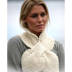 May include: Cream-colored knitted scarf with a textured, chunky knit design. The scarf is wrapped around the neck and chest, creating a layered effect. The scarf appears soft and warm, suitable for cold weather. The background is blurred, suggesting an outdoor setting.