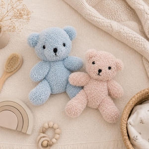 Op de afbeelding: Twee pluche teddyberen, één in lichtblauw en één in zachtroze, met een textuurrijke stof, ronde oren en geborduurde zwarte ogen en neuzen. De beren zijn samen geplaatst en tonen hun pluizige materiaal en eenvoudige, afgeronde vormgeving.