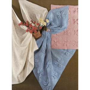 May include: A collection of knitted blankets and shawls in soft pastel colors. A light blue blanket features a floral pattern, while a pink blanket displays a geometric design. A cream-colored shawl with a diamond pattern is also present. A small basket of flowers adds a decorative touch.