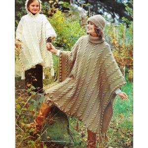 May include: Two people wearing matching knitted ponchos and hats. One poncho is beige, the other is off-white. The beige poncho has a turtleneck and cable knit design. The off-white poncho has a hood and tassels. Both are wearing boots.