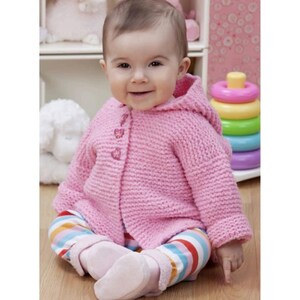 May include: A baby wearing a pink hooded knit sweater with three decorative buttons. The baby is also wearing striped pants with pink socks. A colorful stacking toy is in the background.