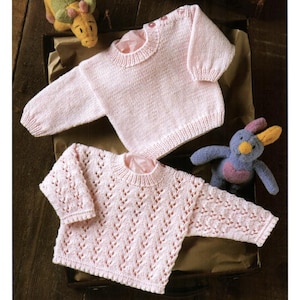 Può includere: Due maglioni rosa chiaro per bambini, uno con una maglia semplice e l'altro con un motivo traforato decorativo. I maglioni sono esposti con due peluche, un dinosauro giallo e verde e un coniglietto blu e rosa.
