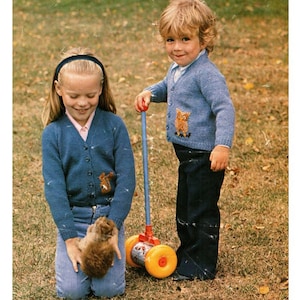 May include: Two children in blue knitted cardigans with animal motifs. The girl holds a small brown animal, while the boy has a red and blue toy. Both wear blue jeans and stand on grass, a classic vintage look.