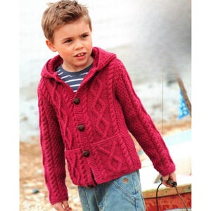 May include: A crimson hooded cable-knit cardigan with three brown buttons and two front pockets. The cardigan is worn over a striped top and blue jeans. The design features a textured cable pattern.