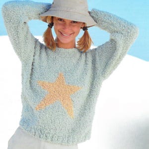 May include: A pale blue sweater featuring a large tan star motif. The sweater has long sleeves and a textured finish. The person in the image is wearing a light-coloured bucket hat.