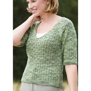 Puede incluir: Un suéter de punto verde claro con cuello en V y mangas cortas. El suéter tiene un diseño texturizado y estampado. La mujer de la imagen lleva una camiseta interior blanca.