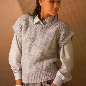 May include: A light grey knitted vest with short sleeves. The vest is worn over a light grey collared shirt with long sleeves. The vest has a ribbed hem and neckline. The shirt sleeves are cuffed at the wrists.