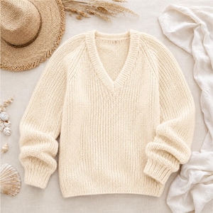 Könnte beinhalten: Cremefarbener V-Ausschnitt-Pullover mit langen Ärmeln. Das Strickwerk hat eine gerippte Textur und eine lockere Passform. Ein Strohhut und Muscheln sind im Hintergrund zu sehen, was auf ein Strand- oder Sommerthema hindeutet.